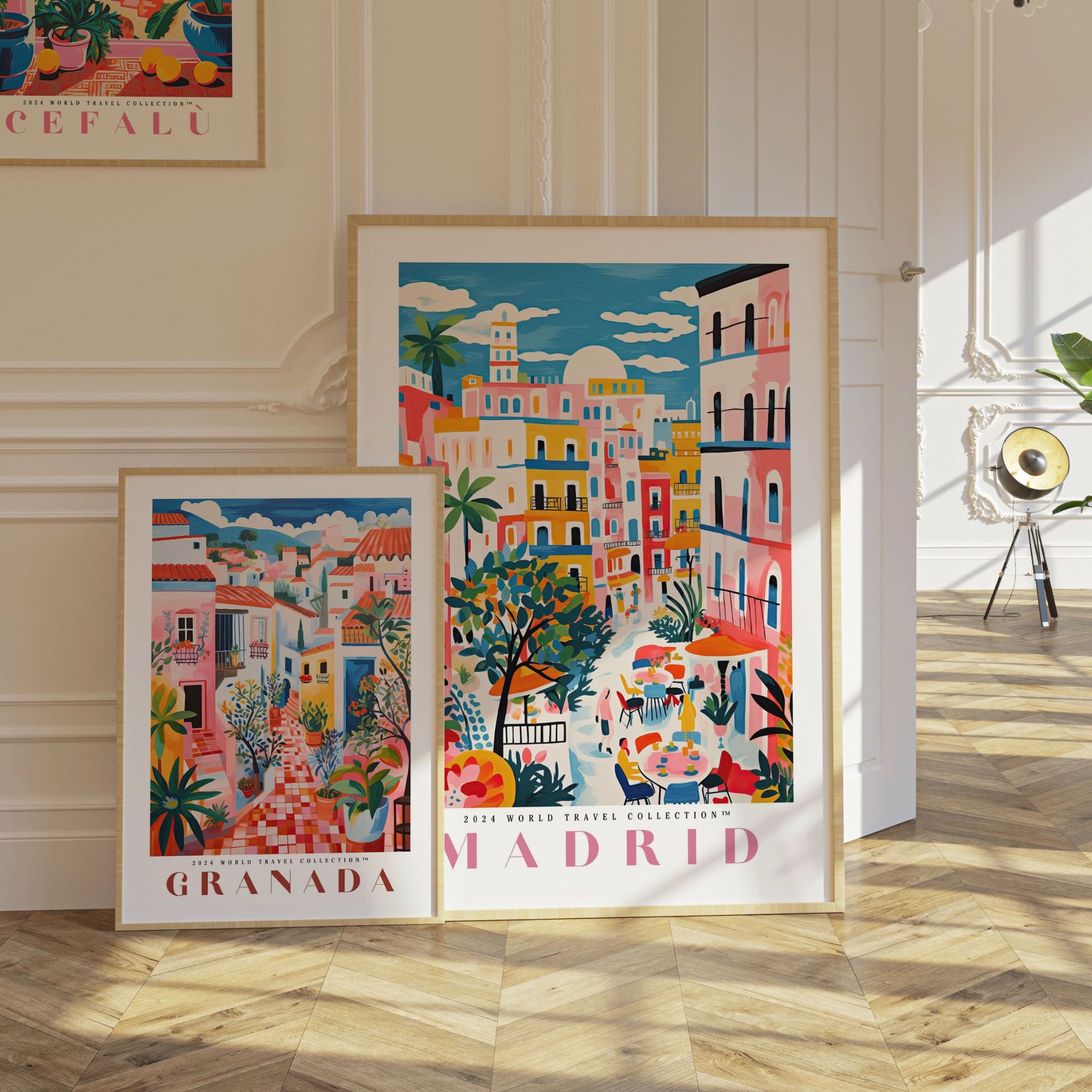 Two framed travel posters featuring vibrant cityscapes: the smaller 'Granada' print with colorful Spanish architecture, lush greenery, and warm red tones, and the larger 'Madrid' print showcasing bold pink, yellow, and blue buildings, lively streets, and outdoor café scenes. Both travel art prints lean against a white-paneled wall on a herringbone wooden floor, surrounded by soft natural light, perfect for adding charm and warmth to any space.