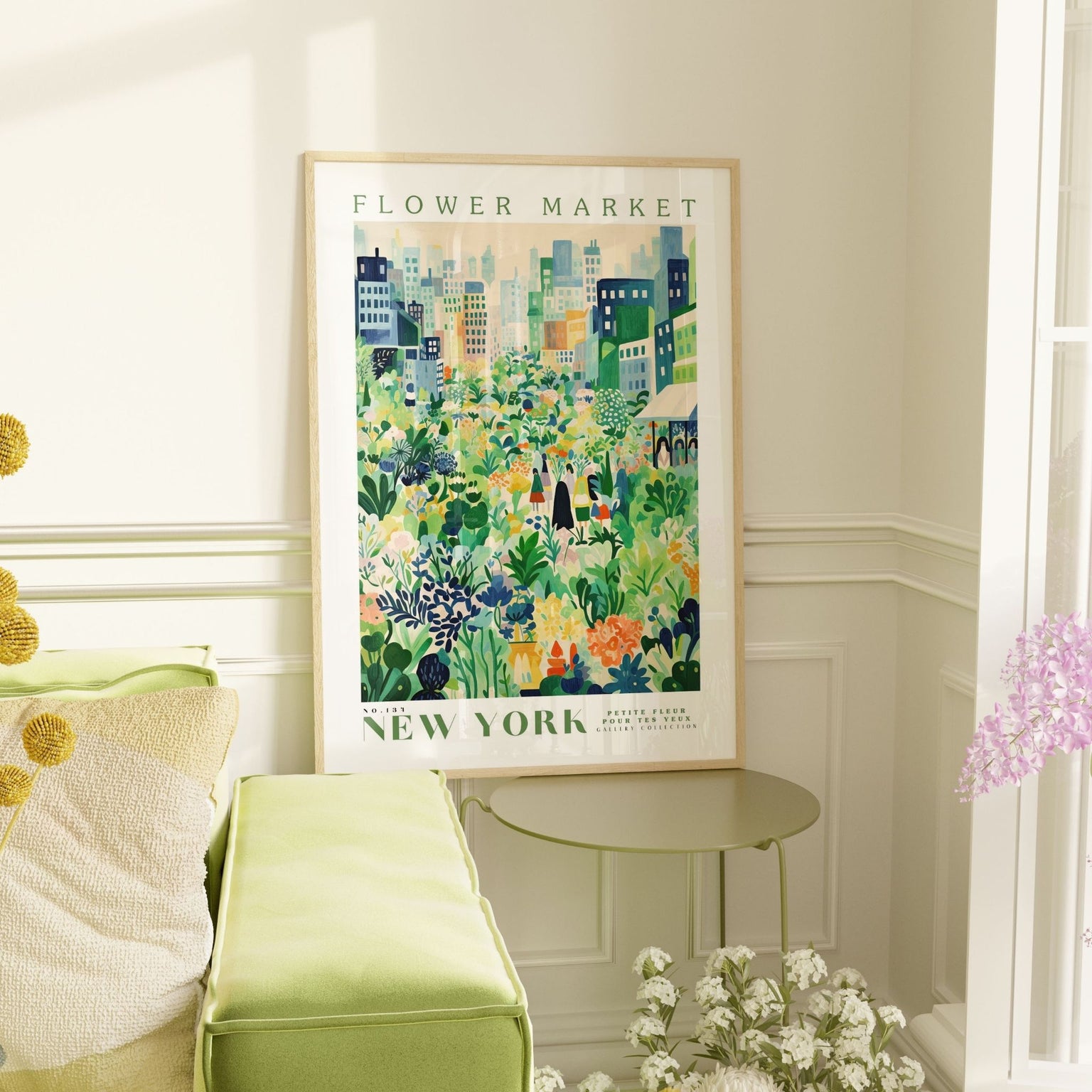 Framed flower market poster titled 'New York' featuring lush green, yellow, and blue floral illustrations set against a cityscape of modern buildings. The poster leans on a soft green bench next to a matching green table, surrounded by white and purple flowers, in a bright and elegant corner filled with natural light.